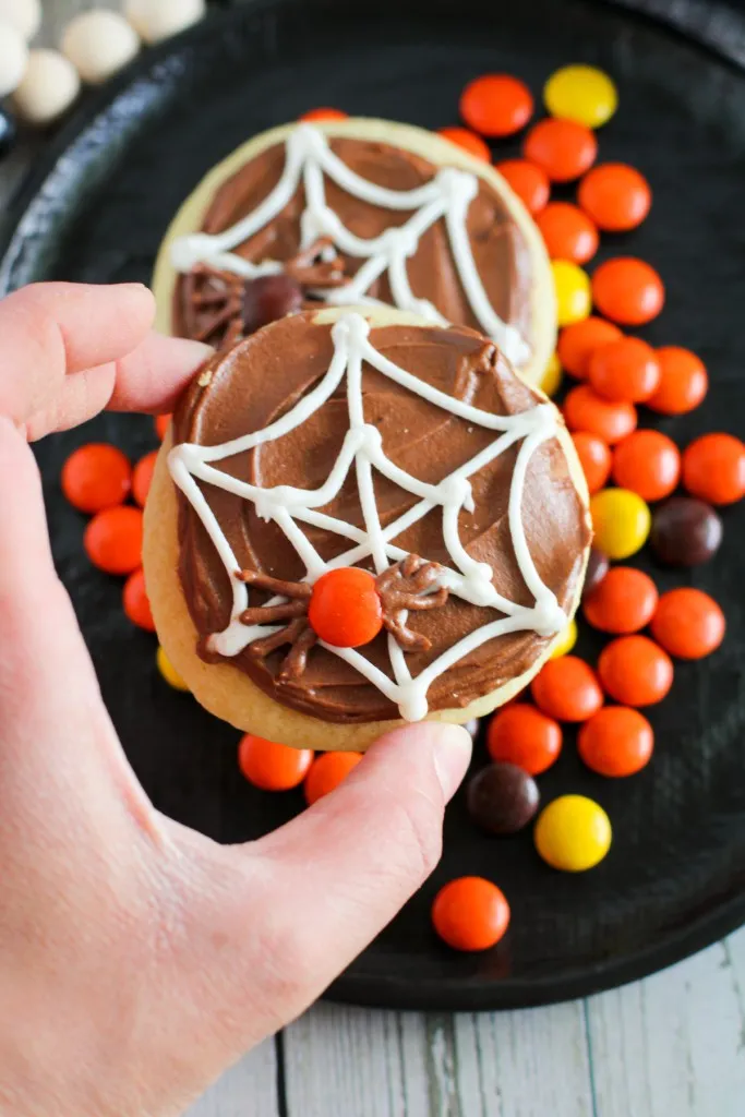Hand holding a spider web sugar cookie above a black plate with additional cookies and Reese's pieces candy.