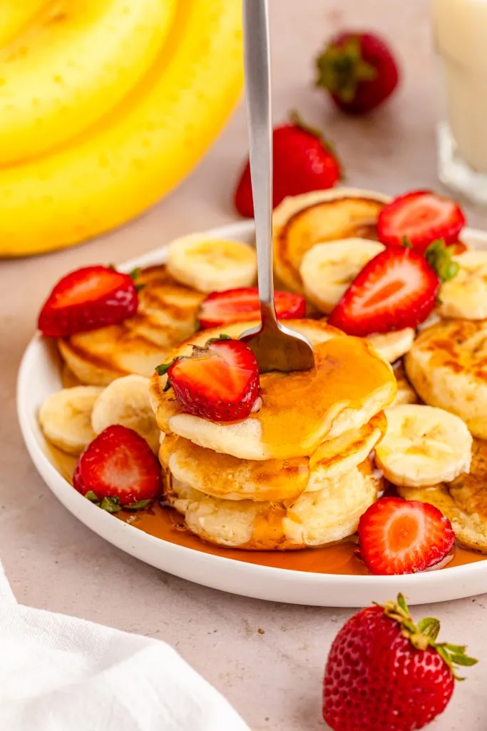 Close up of a plate with mini pancakes, drizzled maple syrup, and freshly slices strawberries and banana.
