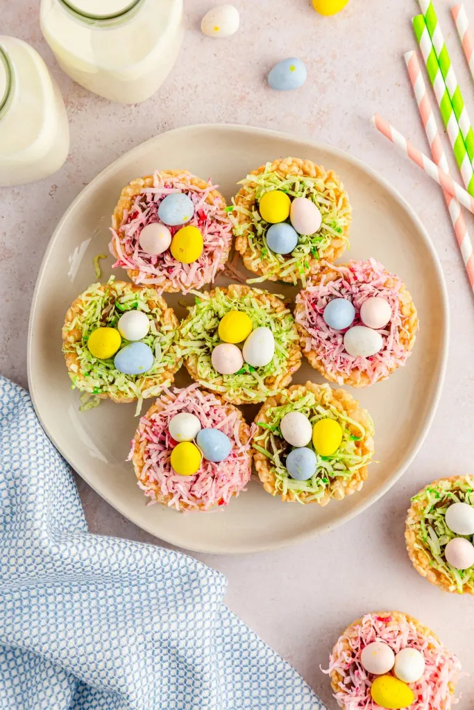 Overhead view of Rice Krispies Easter Nests on a serving plate with jars of milk, pastel blue kitchen cloth, mini eggs candy, and striped straws on a countertop.