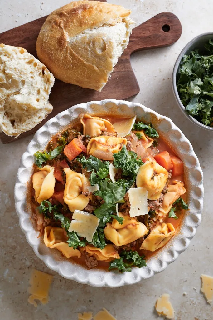 Sausage Kale Tortellini Soup in a white soup bowl, wooden kitchen board with bread, small bowl of kale.