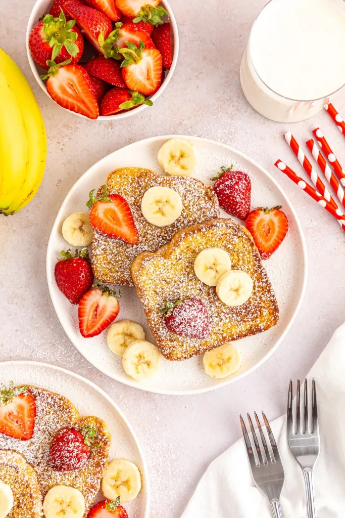 Overhead view of Air Fryer French toast on a white serving plate with powdered sugar, sliced fruit, and maple syrup.