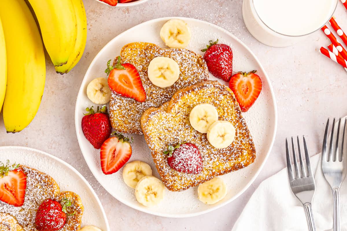 Overhead view of a white serving plate with air fryer French toast with fresh fruit, jug of milk, whole bananas, and metal forks.