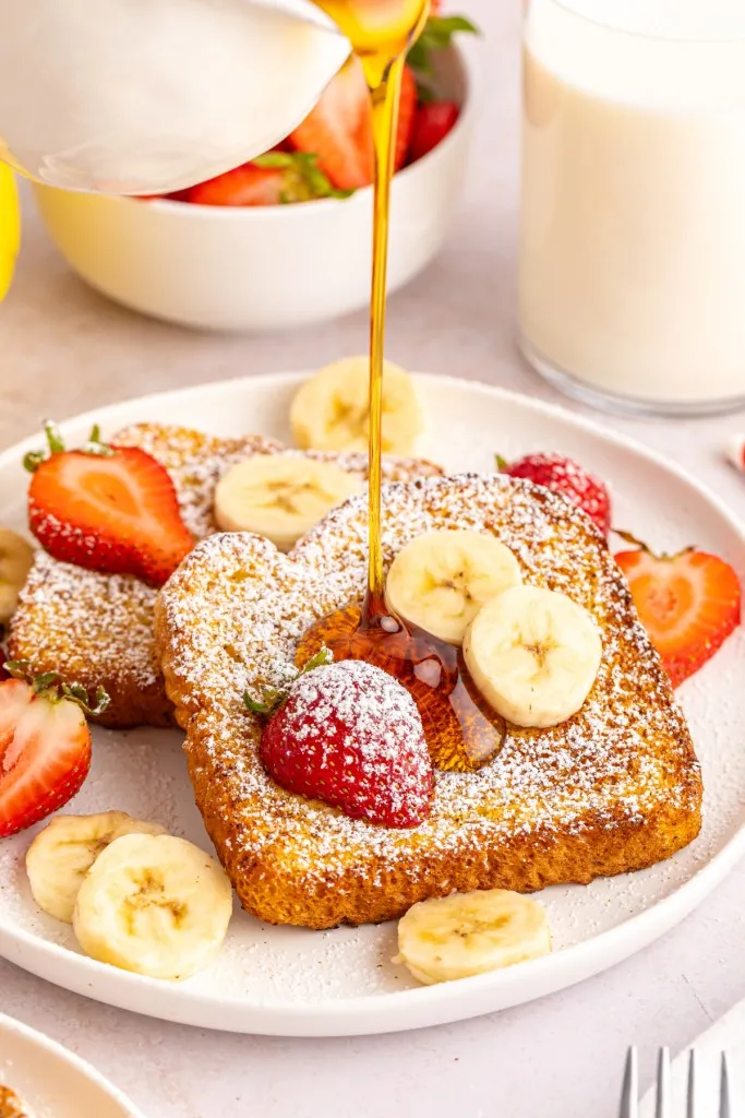 Jug of maple syrup poured over a plate of Air Fryer French toast with sliced fruit and powdered sugar.