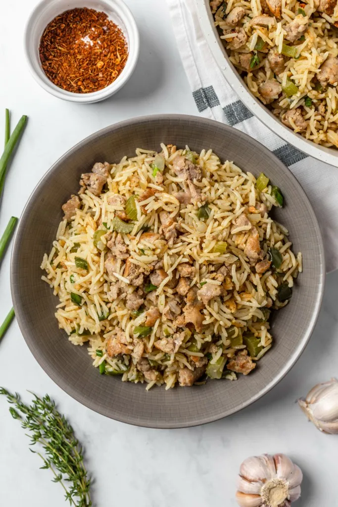 Overhead view of a bowl of dirty rice with sausage, larger bowl with remaining dirty rice with sausage, and individual ingredients scattered around on a white countertop.