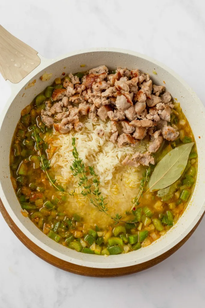 Overhead view of a large skillet with browned sausage, uncooked rice, chicken broth, herbs, and seasoned veggies.