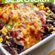 Baked salsa chicken in a white baking dish and wooden serving spoon on a marble countertop.
