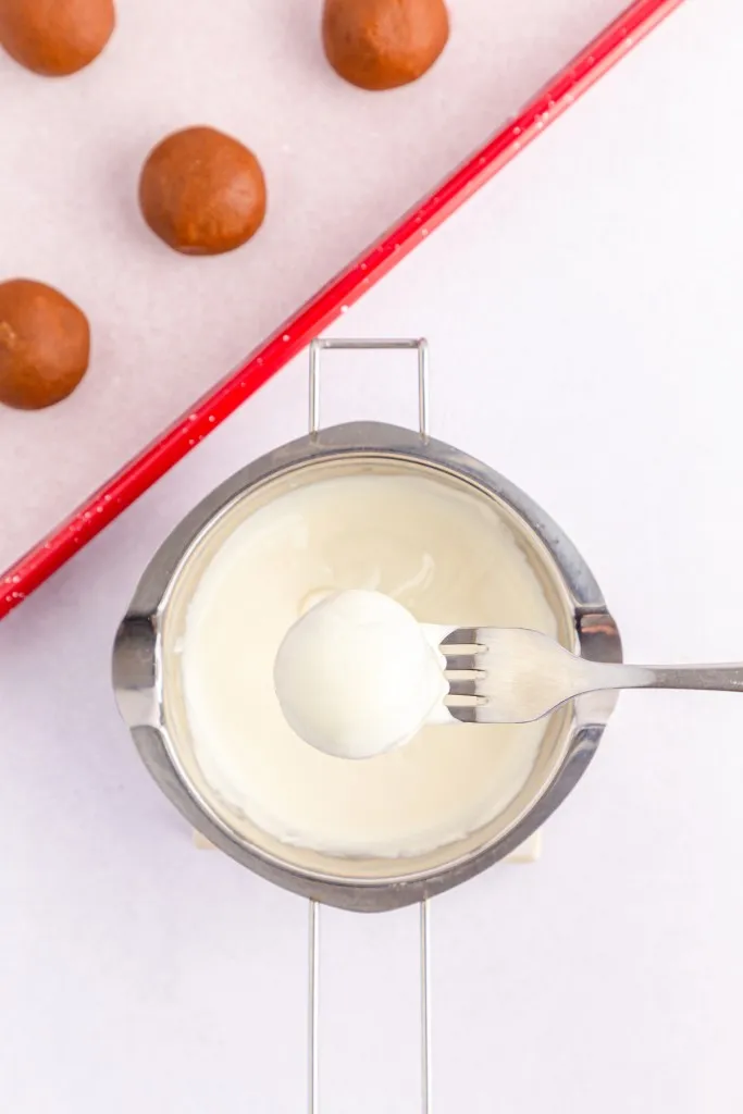 Double boiler with melted vanilla melting wafers, Biscoff ball sitting on a fork that is coated in melted chocolate, tray of chilled Biscoff balls on a baking tray in the background.
