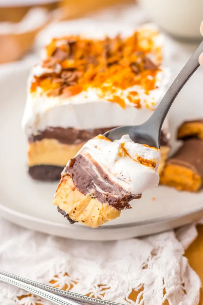 Close up of a spoonful of Butterfinger lush dessert with a slice of Butterfinger lush on a white serving plate with Butterfinger candy.