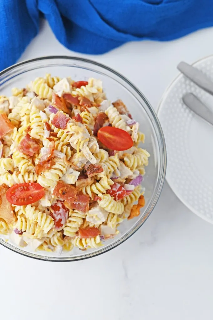 Overhead view of a bowl of chicken bacon ranch pasta salad on a marble countertop with a blue kitchen cloth in the background, white serving plate with metal utensils to the side.