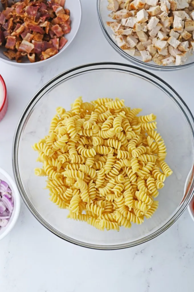 Overhead view of the cooked al dente rotini pasta in a glass bowl, surrounded by the other prepared pasta salad ingredients.