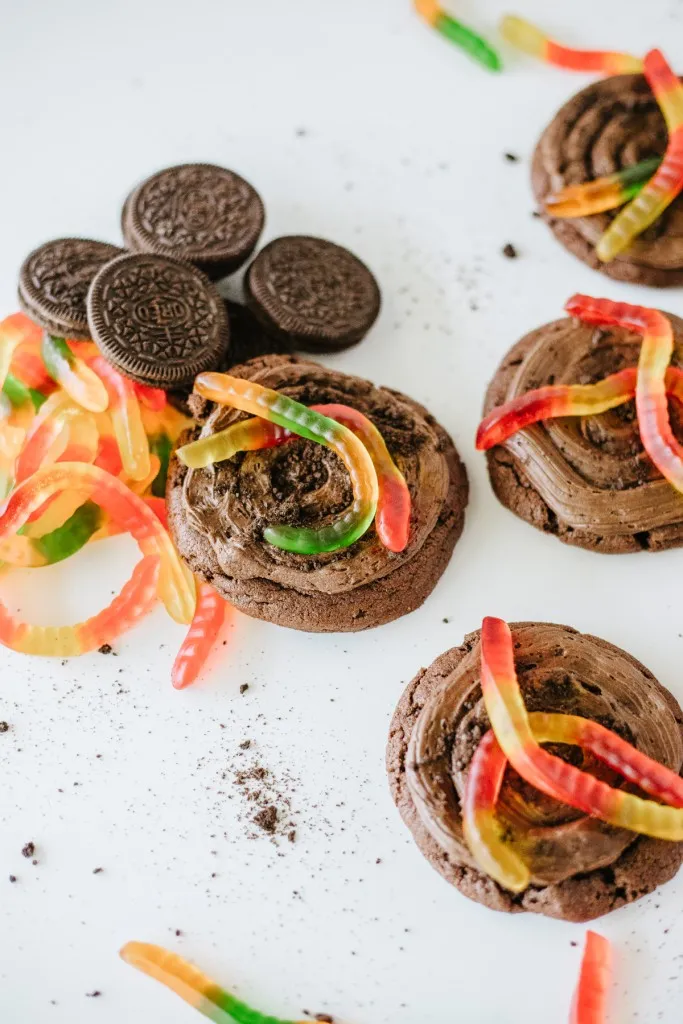 Stack of copycat Crumbl dirt cake cookies on a white counter top with oreo crumbles and gummy worms around