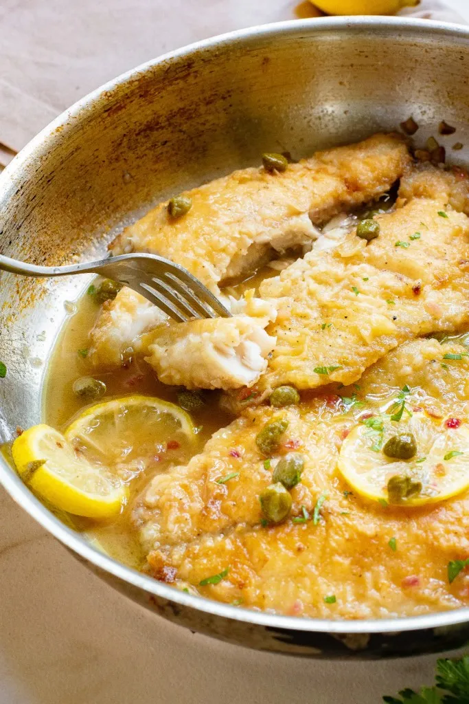Close up of a large frying pan with Tilapia Piccata, metal fork piercing a piece of fish.