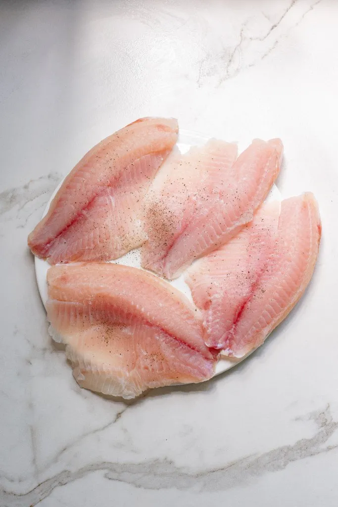 Fresh Tilapia fillets seasoned with sea salt and ground black pepper on a white plate.