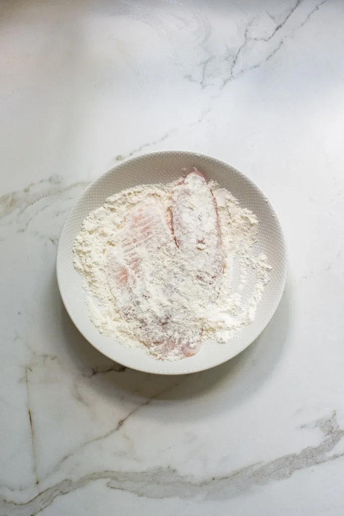 Seasoned Tilapia fish dredged in flour on a white plate.