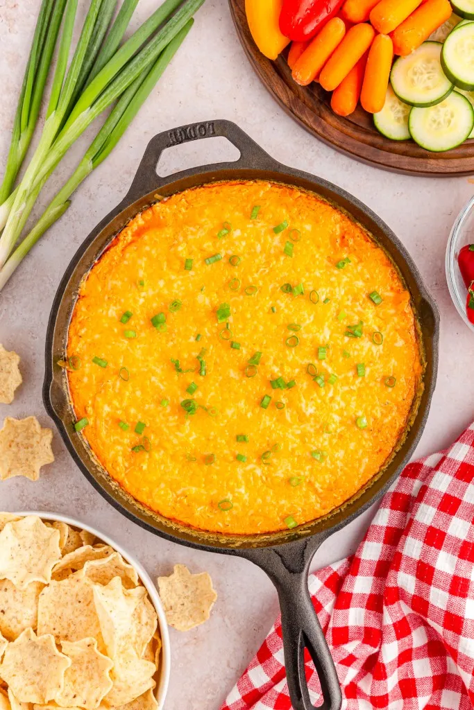 Overhead view of a skillet filled with baked buffalo chicken dip, freshly sliced veggies, and a bowl of crackers and tortilla chips.