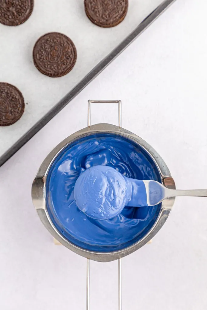 Overhead view of double boiler with melted blue candy melts, Oreo cookie fully coated in the melted chocolate and resting on a metal fork.