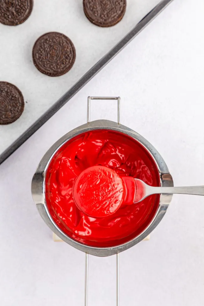 Overhead view of double boiler with melted red candy melts, Oreo cookie fully coated in the melted chocolate and resting on a metal fork.