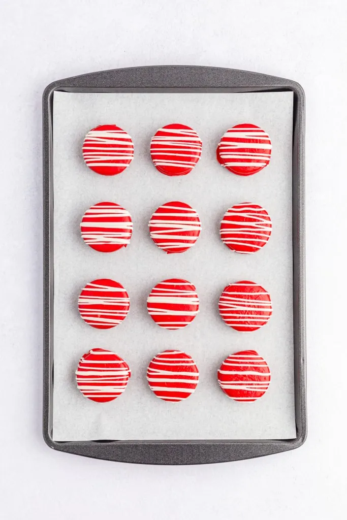 Overview of a baking tray lined with parchment paper and Oreo cookies that have been coated in melted red candy melts with a drizzle of white melted chocolate.