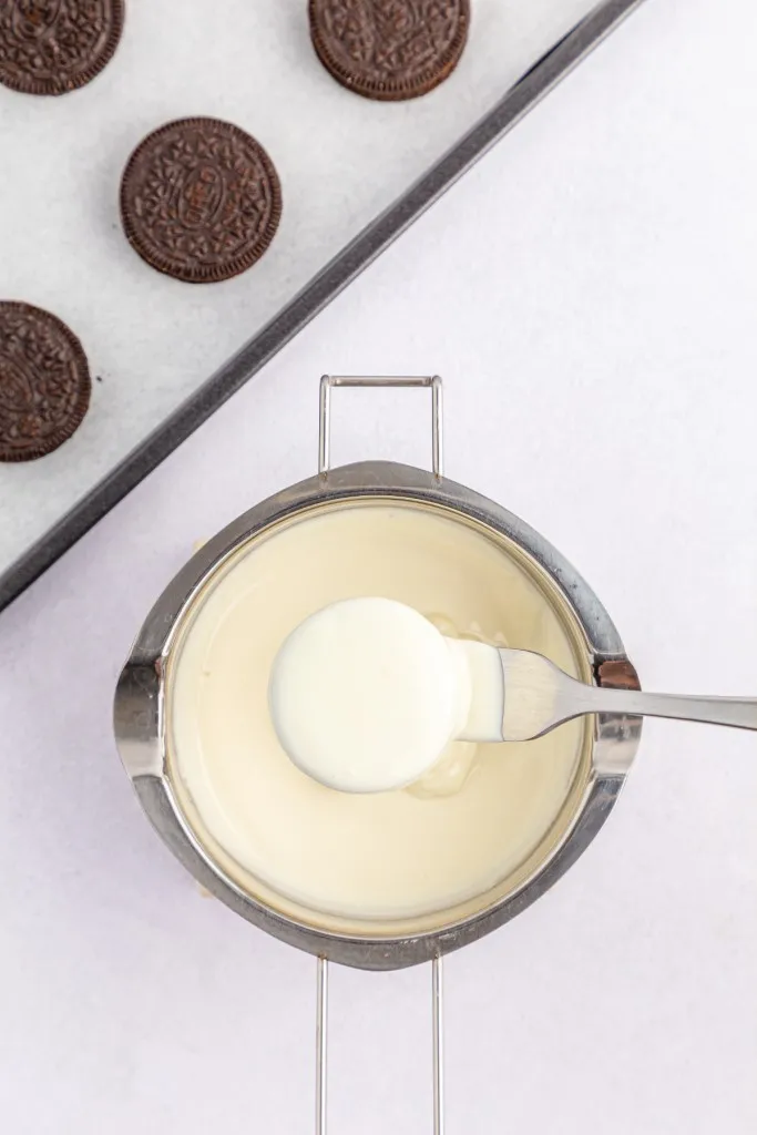 Overhead view of double boiler with melted white wafers, Oreo cookie fully coated in the melted chocolate and resting on a metal fork.