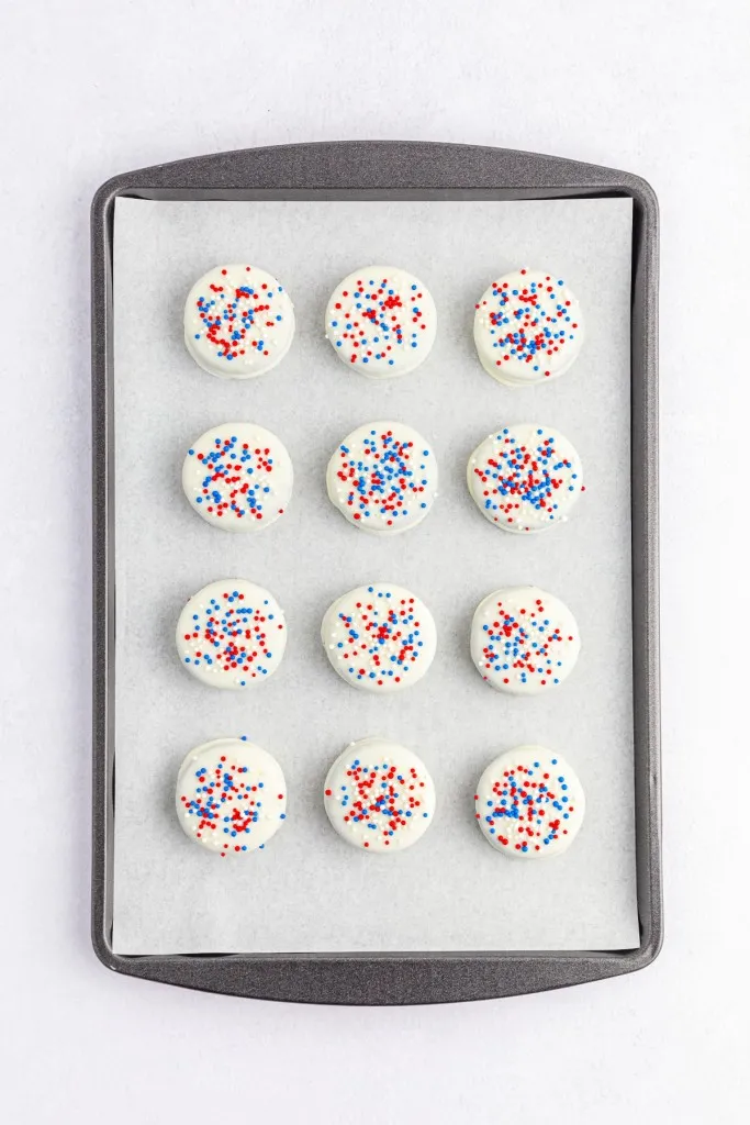 Overview of a baking tray lined with parchment paper and Oreo cookies that have been sprinkled with red, white, and blue nonpareils.