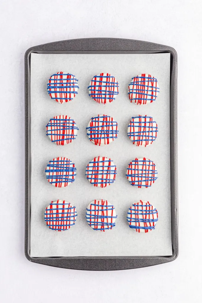 Overview of a baking tray lined with parchment paper and Oreo cookies that have been coated in melted white chocolate with a drizzle of red and blue melted chocolate.