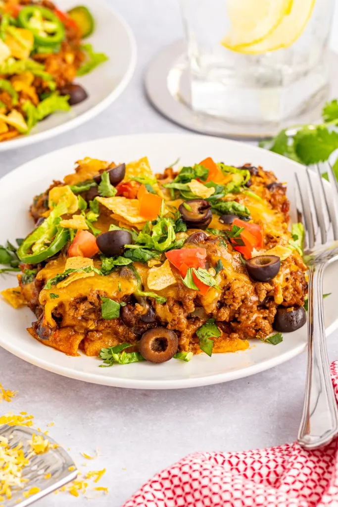 Close up of a serving of easy taco casserole on a white plate with a silver fork, a glass of water and second plated serving in the background.