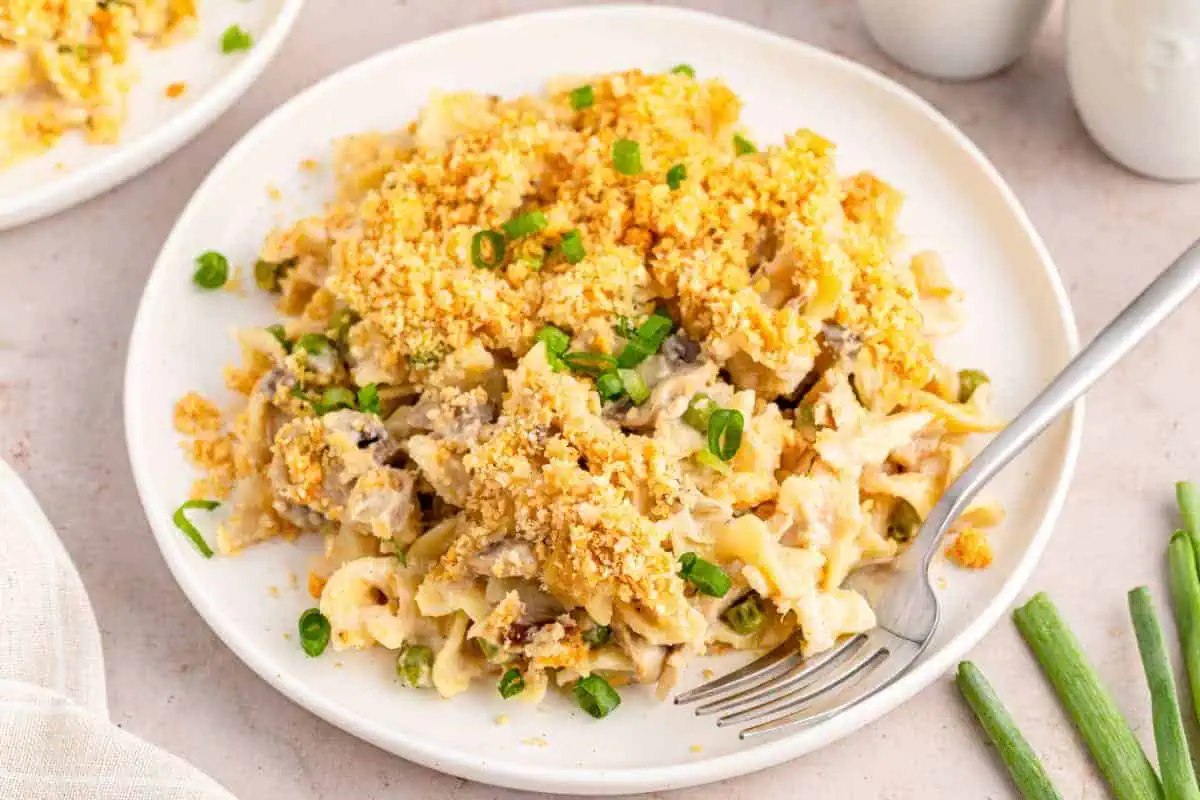 A serving of tuna noodle casserole on a white serving plate with a silver fork, green onions and a white kitchen cloth in the background.