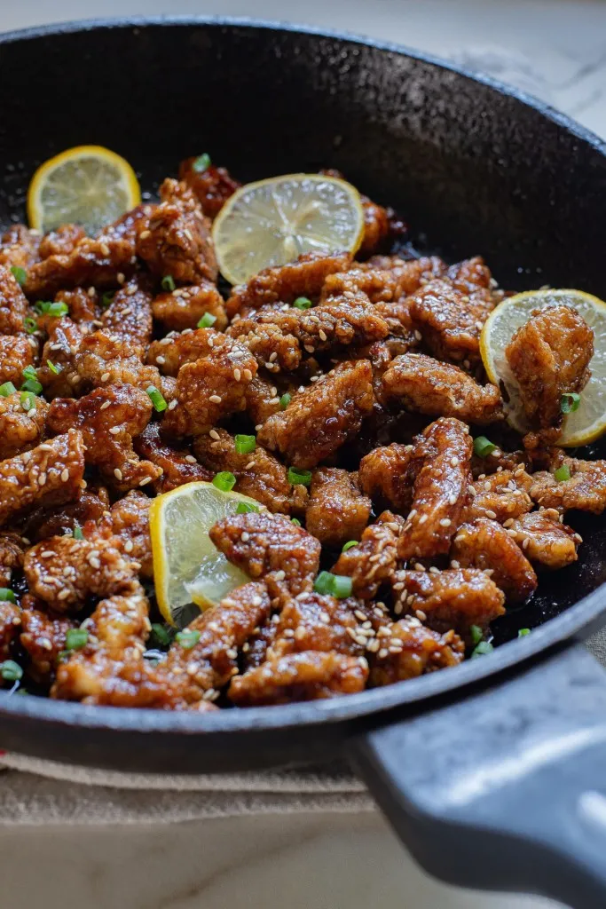 Close up of Chinese lemon chicken in a skillet with sesame seeds, lemon slices, and chopped green onions.