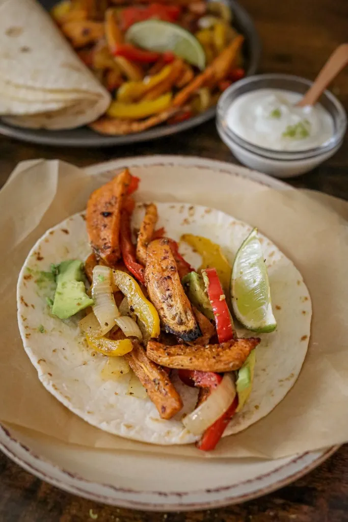 Sweet potato fajita filling loaded onto a flour tortilla, small bowl of lime sauce, sliced avocado, and baking tray with roasted fajita veggies and heated tortillas in the background.