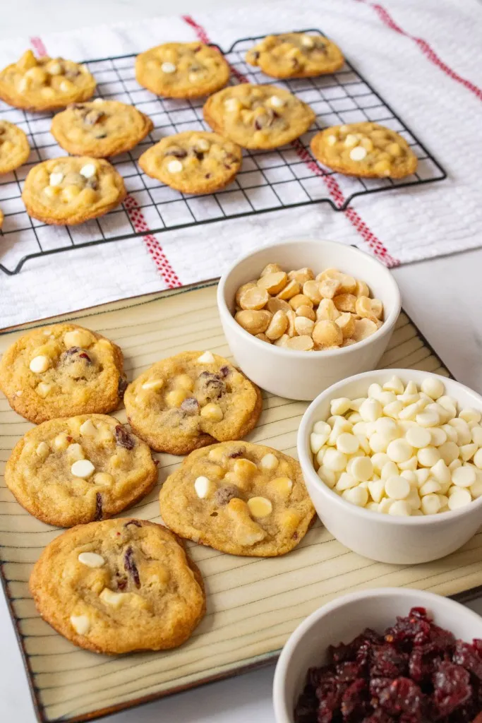 White chocolate cranberry hazelnut cookies on a serving plate with white ramekins filled with nuts, white chocolate chips, and dried cranberries. Cookies in the background cooling on a wire rack.
