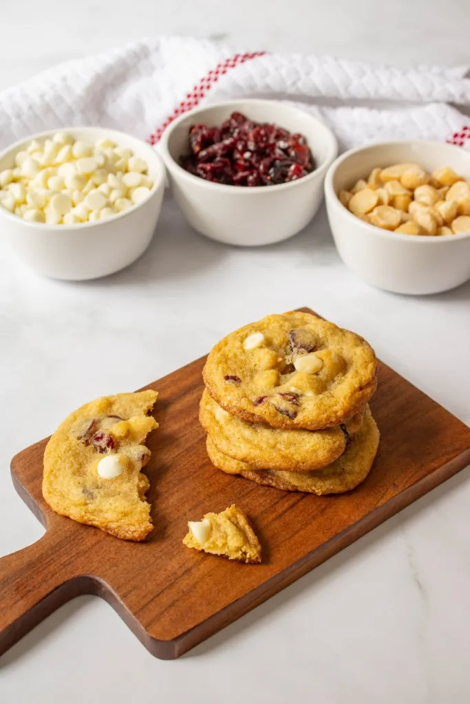 White chocolate cranberry hazelnut cookies stacked on a wooden kitchen board, white ramekins with white chocolate chips, dried cranberries, and nuts.