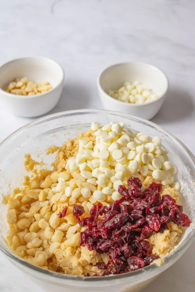 Glass mixing bowl with cookie dough, white chocolate chips, nuts, and dried cranberries. White ramekins with nuts white chocolate chips.