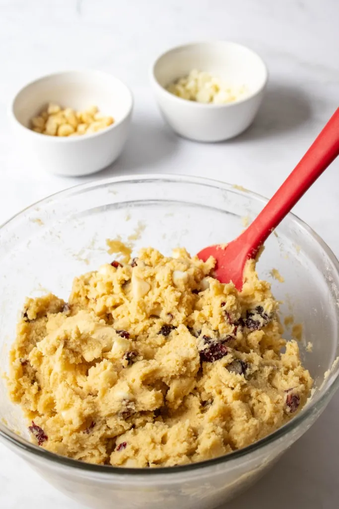 Glass mixing bowl with cookie dough, white chocolate chips, nuts, and dried cranberries with plastic spatula. White ramekins in the background with white chocolate chips and nuts.