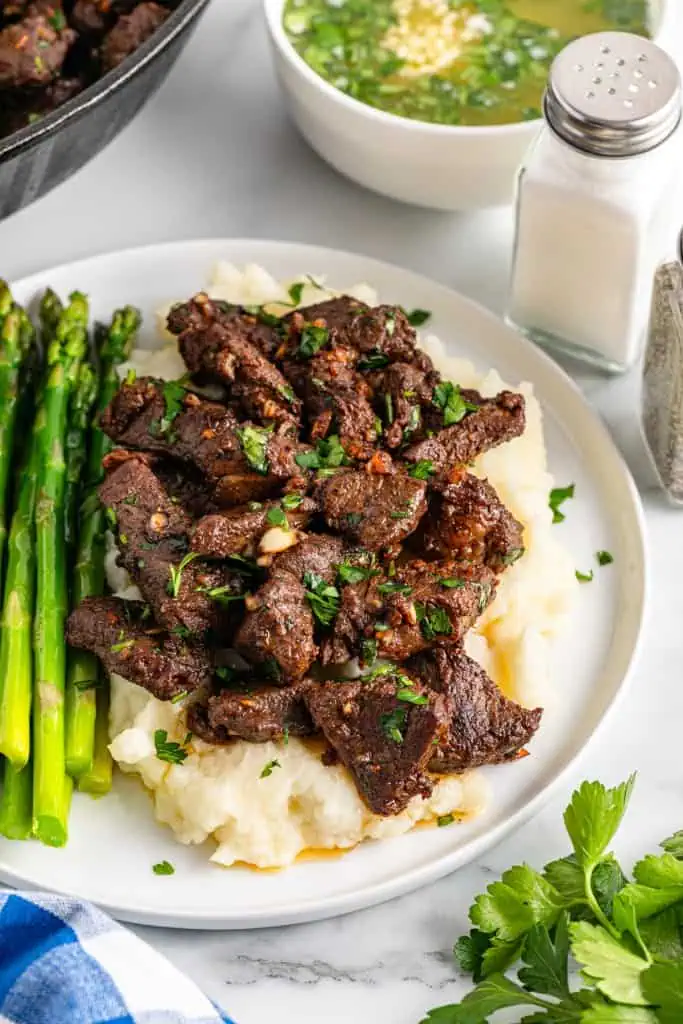 Garlic butter steak bites topping mashed potato and sitting alongside roasted asparagus on a white serving plate.