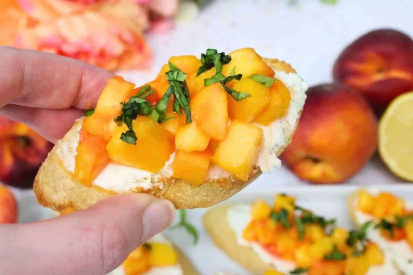 a hand holding a slice of bruschetta topped with peach chunks and goat cheese.