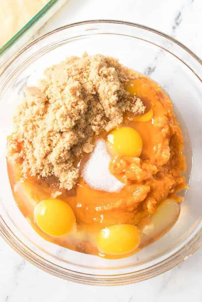 A large glass mixing bowl with canned pumpkin, eggs, salt, and brown sugar.
