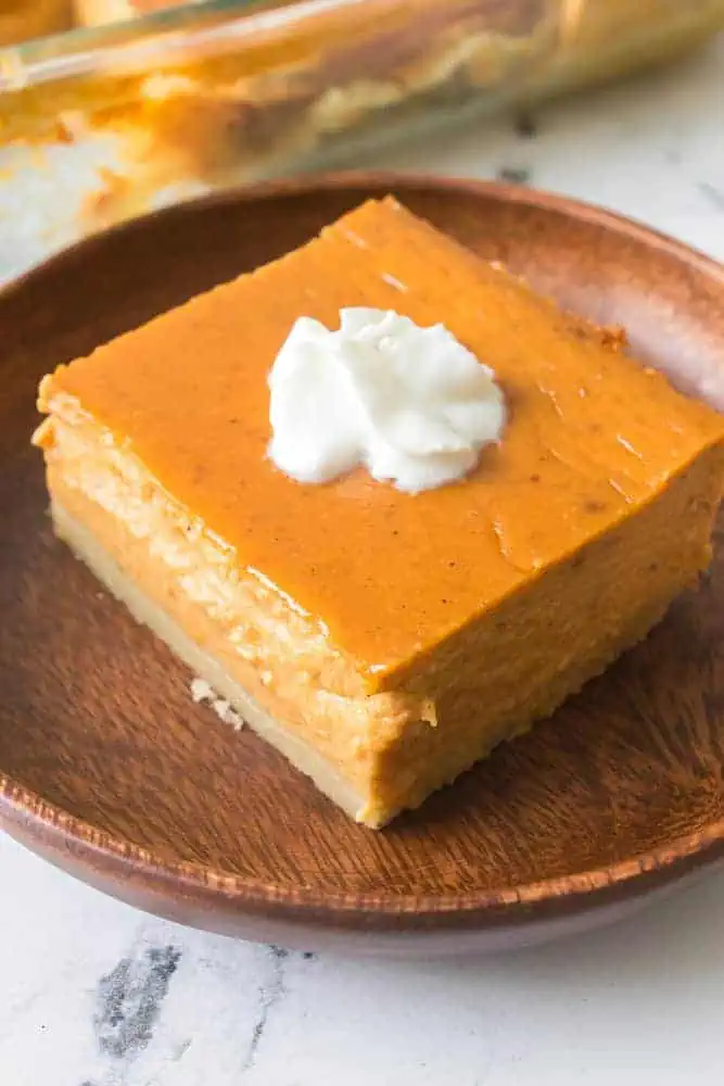 Close up of a pumpkin pie dessert bar, topped with a small dollop of whipped cream, sitting on a wooden plate.