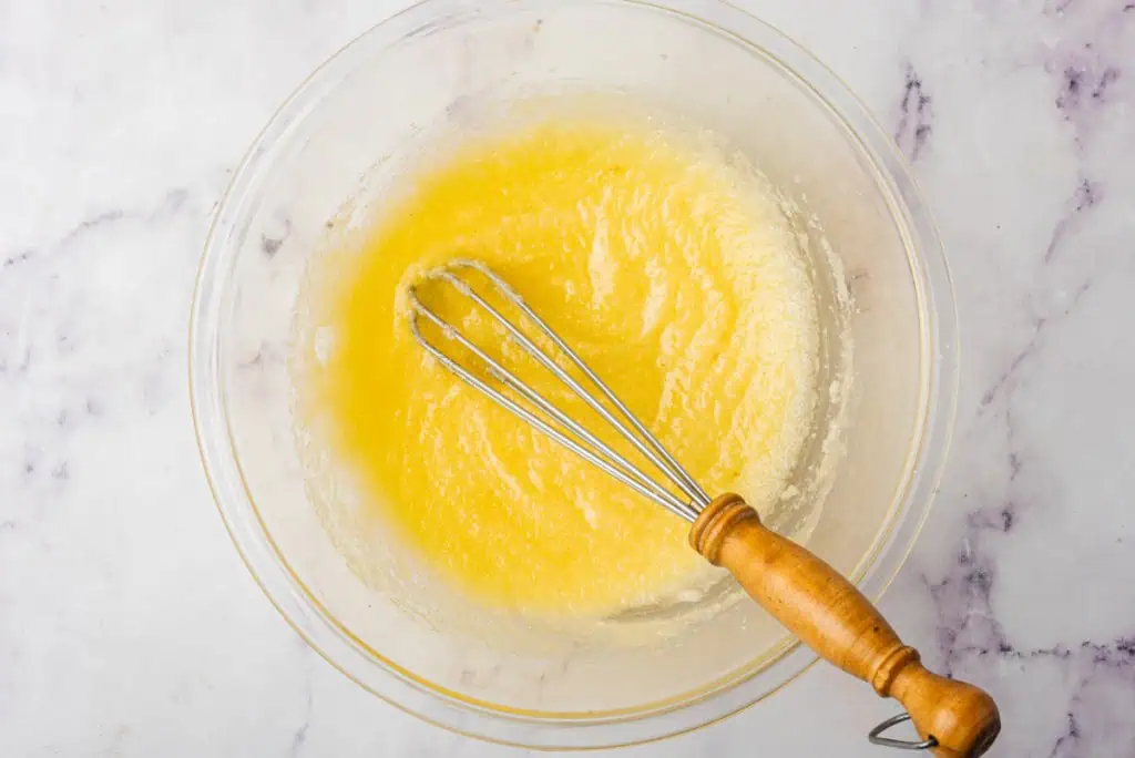 Glass mixing bowl with melted butter and granulated sugar with a small metal whisk.