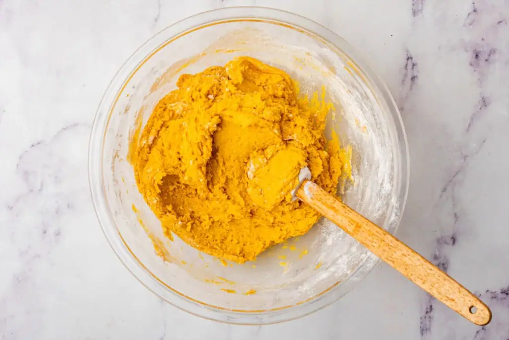 Glass mixing bowl with pumpkin puree cookie mixture, spices, flour, baking soda, baking powder, and salt with a plastic spatula.