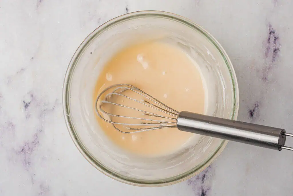 Small mixing bowl with powdered sugar and milk and a metal whisk.