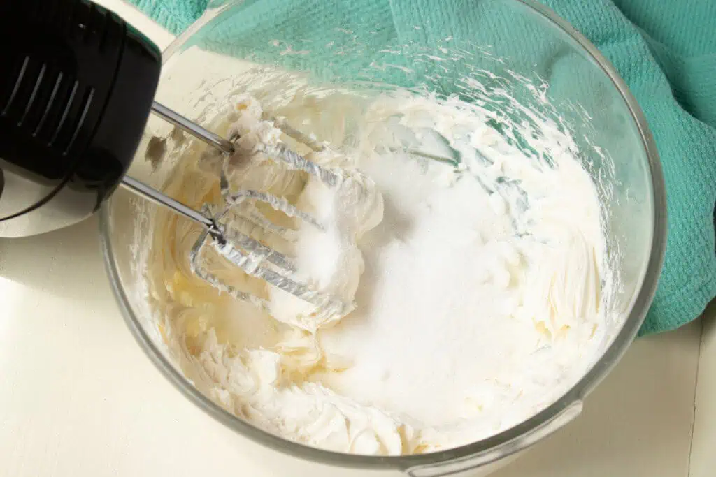 Glass bowl with softened cream cheese and granulated sugar, electric mixer with attachments covered in cream cheese mixture.