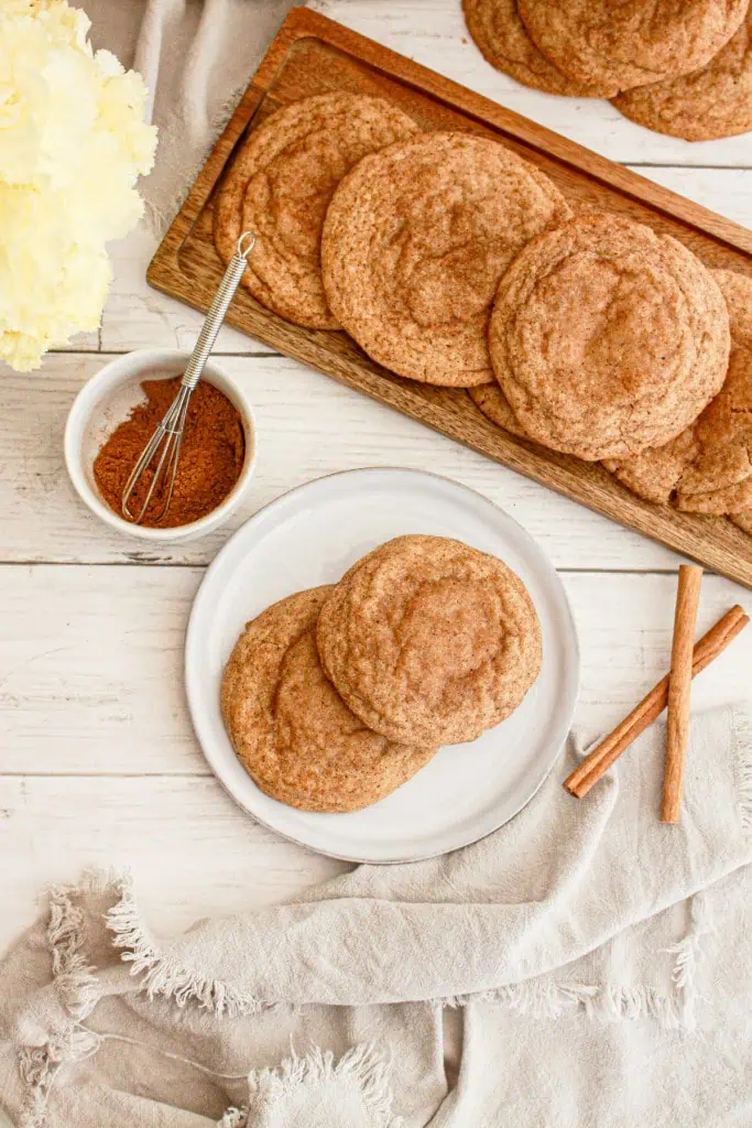 Two pumpkin cheesecake cookies on a white serving plate, additional cookies on a long wooden serving board, small bowl with pumpkin spice and a small metal whisk, cinnamon sticks on the wooden table along with a cream kitchen cloth.
