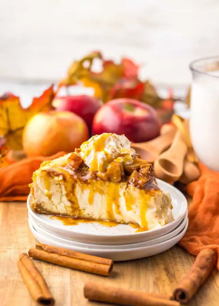 A slice of caramel apple cheesecake on stacked white side plates, topped with softened apples, whipped cream, and caramel drizzle.