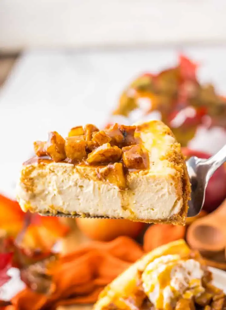 Close up of a slice of caramel apple cheesecake on a silver server, topped with apple topping and caramel drizzle.
