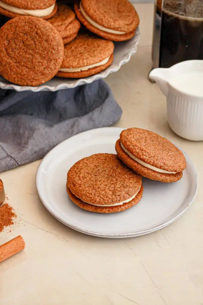 Two gingerbread latter sandwich cookies on a white serving plate, stacked gingerbread latter sandwich cookies on a cake stand in the background, small jug of milk.