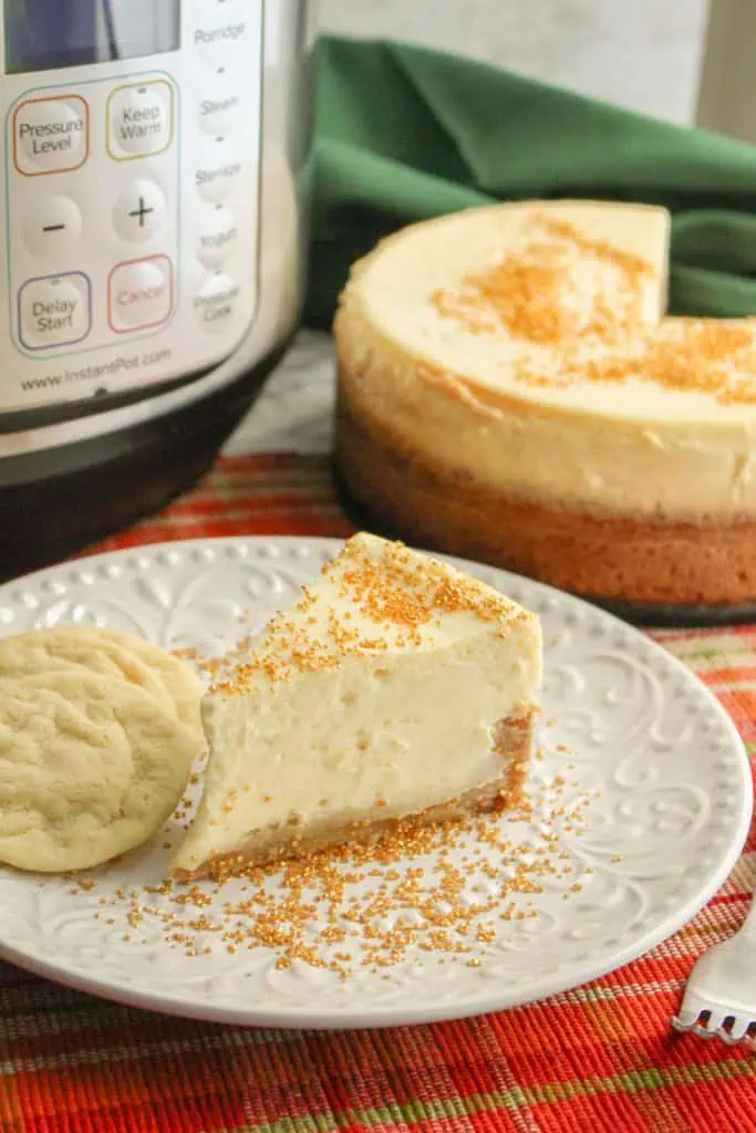 Slice of Instant Pot Sugar Cookie Cheesecake with three sugar cookies on a white serving plate with the remainder of the cheesecake and Instant Pot appliance in the background.