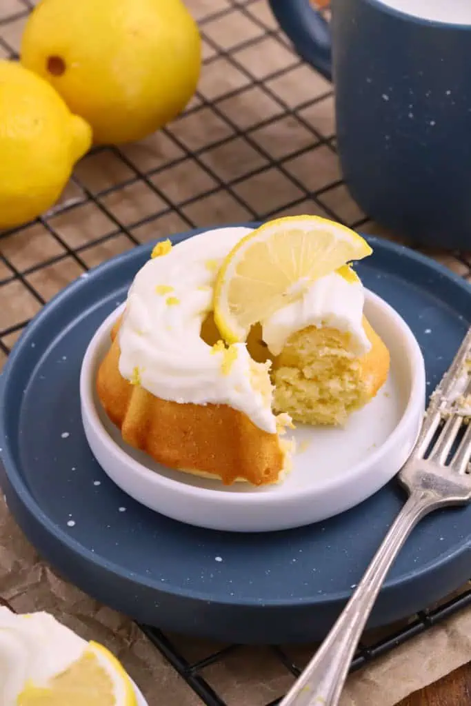 Mini Lemon Bundt Cake on a blue serving plate with a forkful removed from the cake, fork resting beside the cake and fresh lemons in the background.