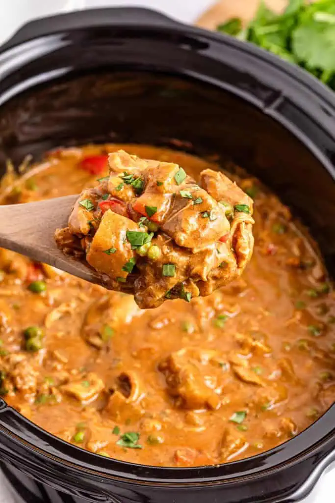 A wooden spoonful of Slow Cooker Chicken Curry held above the remainder of the curry in the slow cooker pot.
