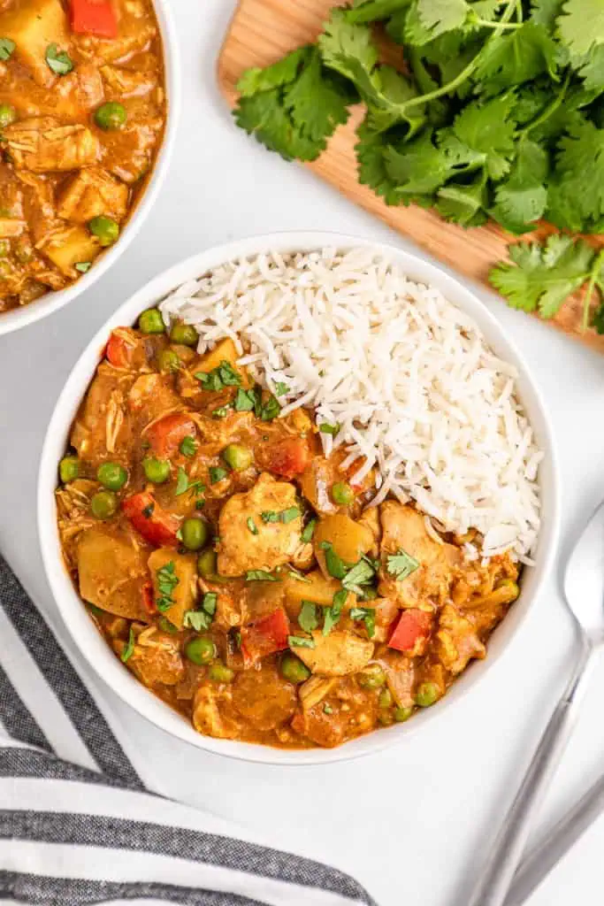 Overhead view of a bowl of Slow Cooker Chicken Curry with rice, striped kitchen cloth, silver spoon, extra bowl of curry, and fresh parsley scattered around.