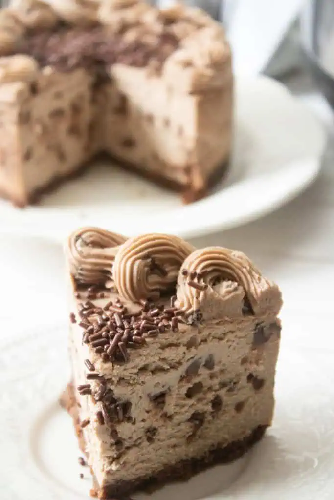 Close up of a slice of Instant Pot Chocolate Chip Cheesecake on a white plate with the remainder of the cheesecake in the background.